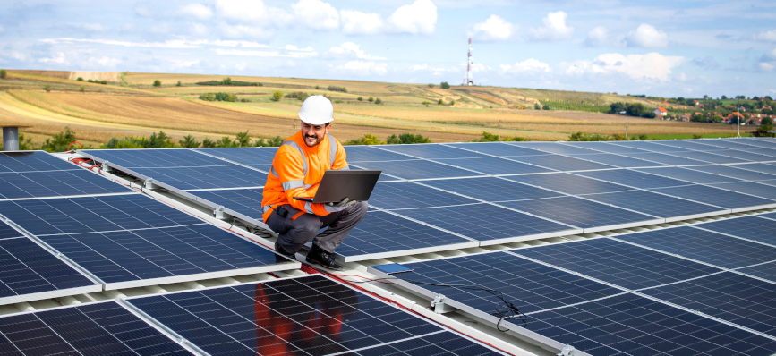 A man wearing a hard hat and high visibility jacket holds a laptop while standing on a roof covered with solar panels.
