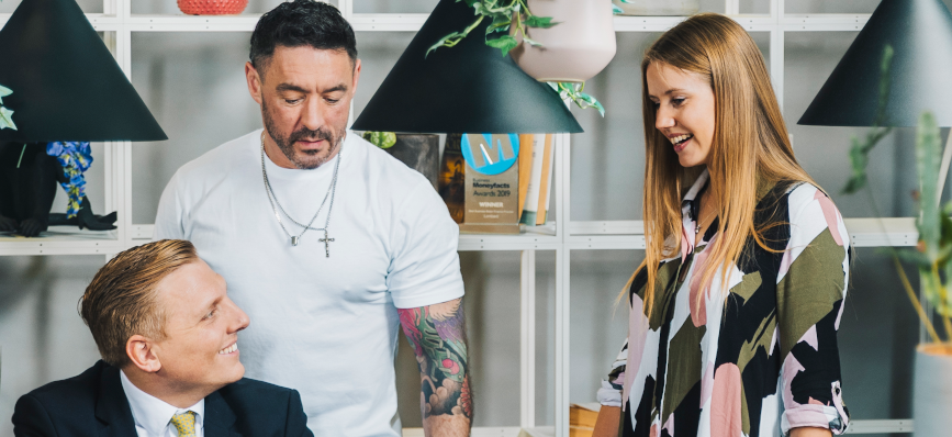 This image shows three people together. A man wearing a tie is sat down and looking at a women who is standing up. A man in white t-shirt stands alongside them both.