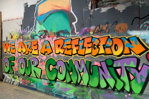 Graffiti mural reads ‘WE ARE A REFLECTION OF OUR COMMUNITY’ in bold orange, green, and purple letters on a colorful wall, linked to Cardiff-based non-profit social enterprise Media Academy