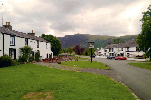 Quiet village in Cumbria with white cottages, a pub sign, green lawns, and hills in the background under cloudy skies.