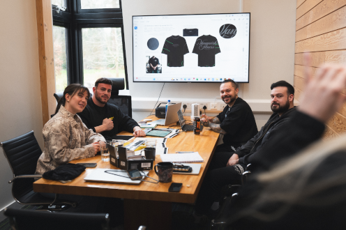 Meeting room with people at Vanquish Fitness around a wooden table, laptops and notebooks, and a screen showing black t-shirt designs.