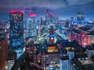 Nighttime cityscape of Manchester featuring the illuminated Central Library clock tower, surrounded by modern high-rises and vibrant city lights.