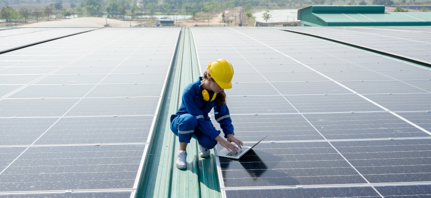 A woman crouching on a roof covered by solar panels types on a laptop