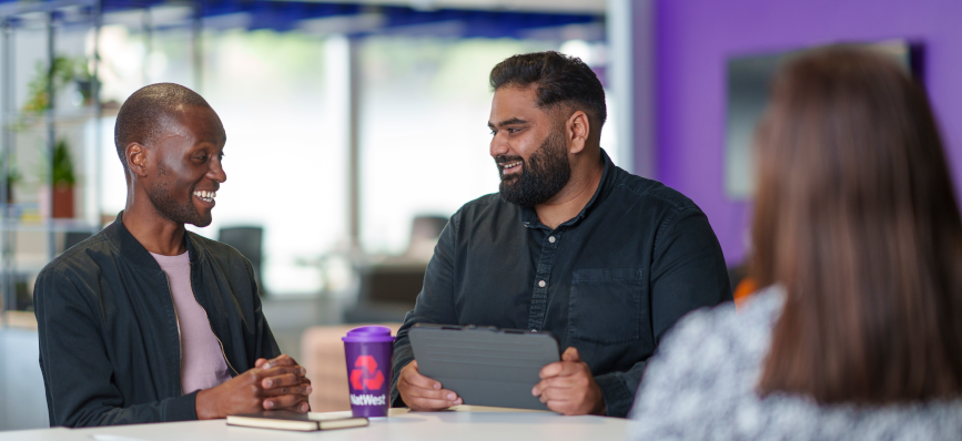 Two men sitting at a table smiling at one another in a business meeting setting, with one holding a tablet. A NatWest branded purple cup is between them on the table.