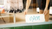 A lady holds and 'open' sign at the window of a dress shop