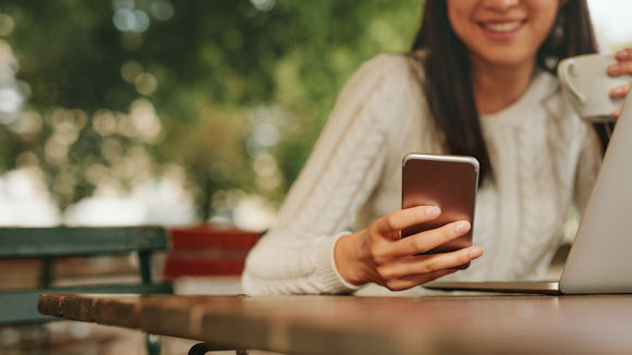 Young woman in a cafe reading a text message from her mobile phone. Chinese female sitting at cafe table with laptop and using smart phone.