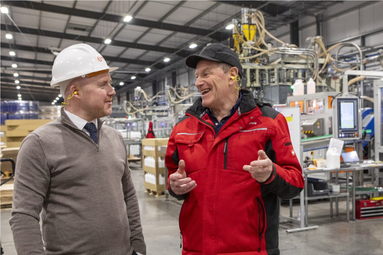 Two people wearing safety gear stand in a manufacturing facility, discussing operations. One person is wearing a hard hat and business attire, while the other is dressed in a red work jacket. Industrial machinery, piping, and production equipment fill the background.