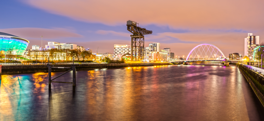Nighttime view of Glasgow’s River Clyde with reflections of city lights, a green-lit building, old crane, and purple-lit arch bridge.